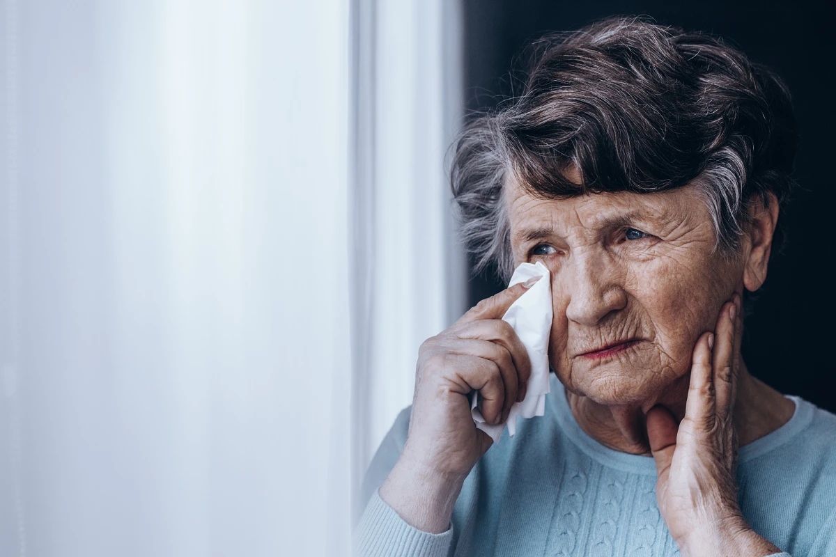 an elderly woman with watery eyes wiping her eye with a tissue