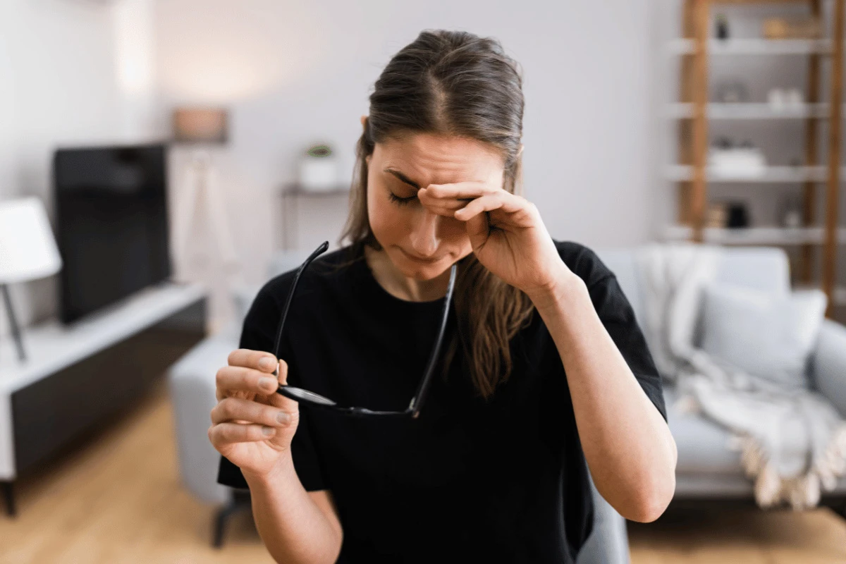 a woman sitting at home taking off her glasses and rubbing her eye
