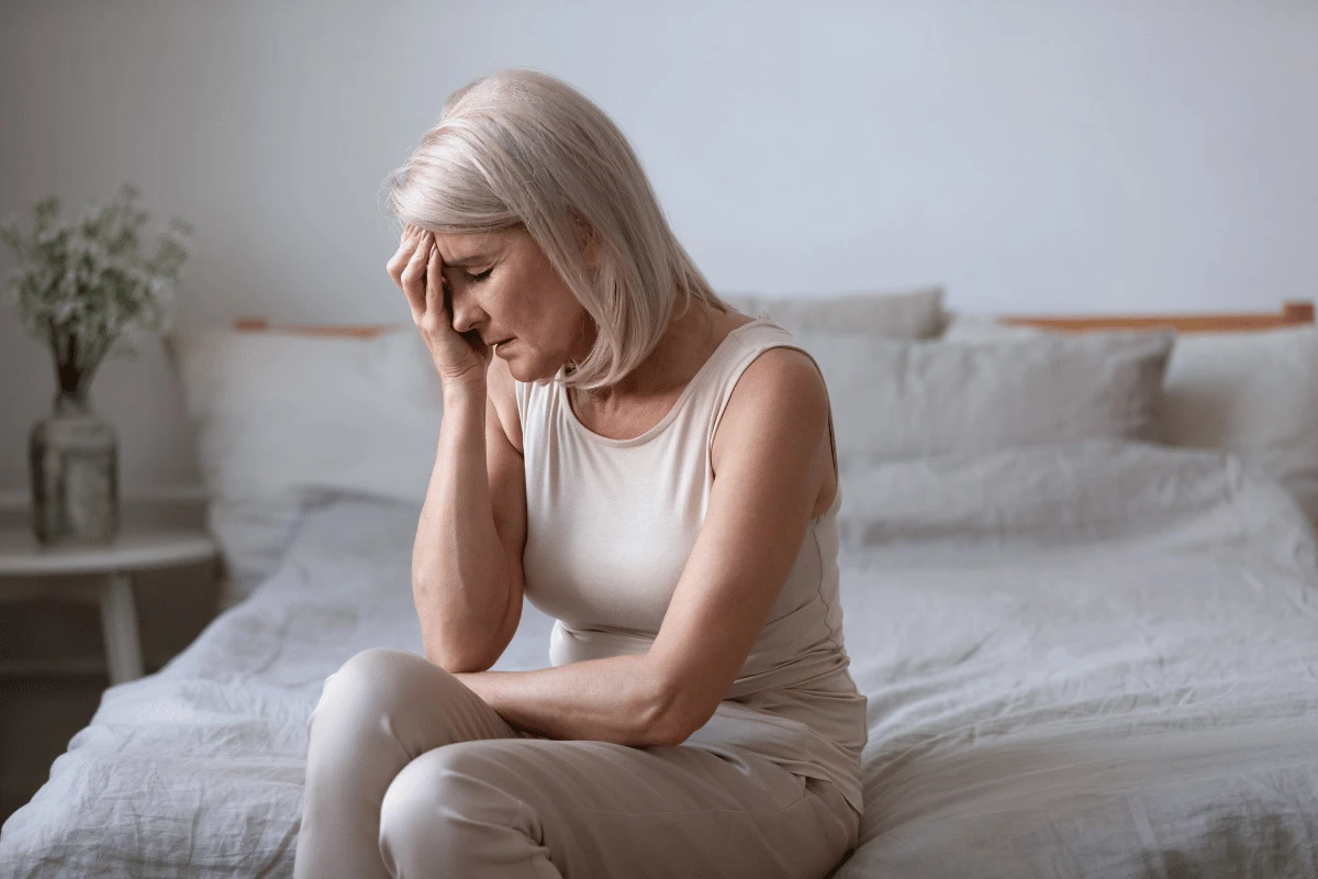 an older woman sitting on a bed and holding a hand to her head