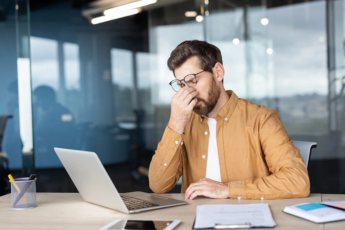 a man sitting at his desk at work looking uncomfortable and massaging the bridge of his nose