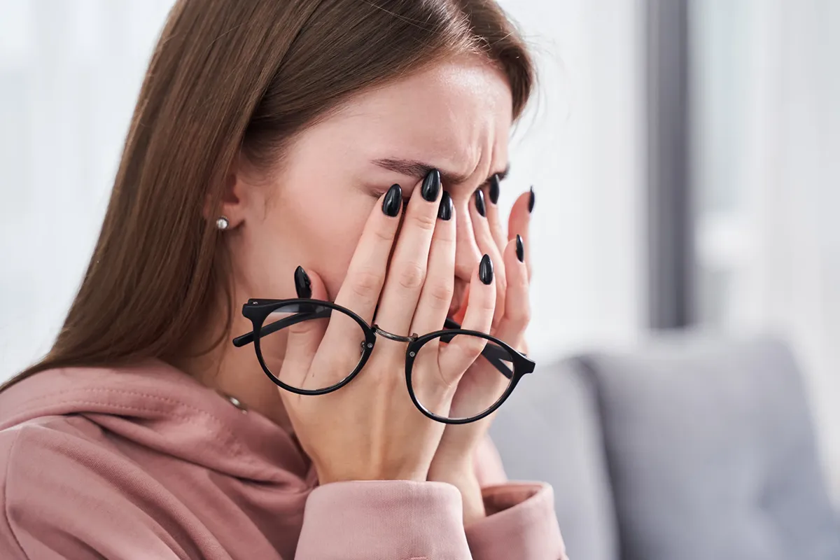 An anxious young woman holds her glasses and rubs her eyes.