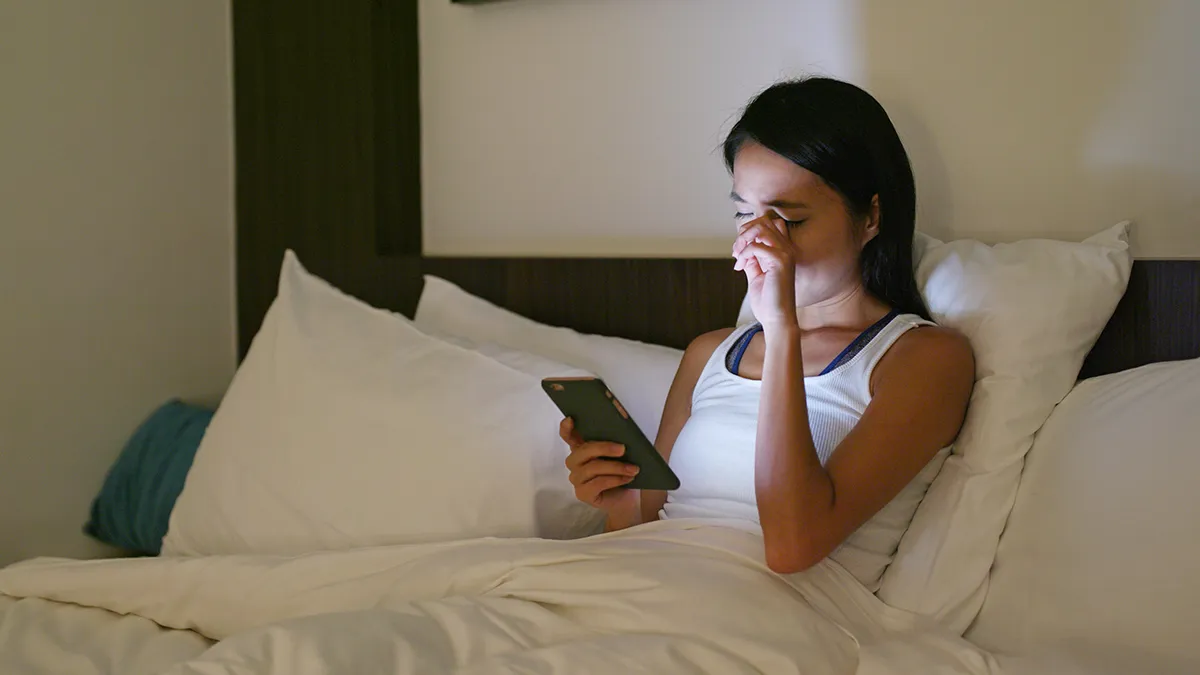 a woman in bed at night looking at her phone and rubbing her eye