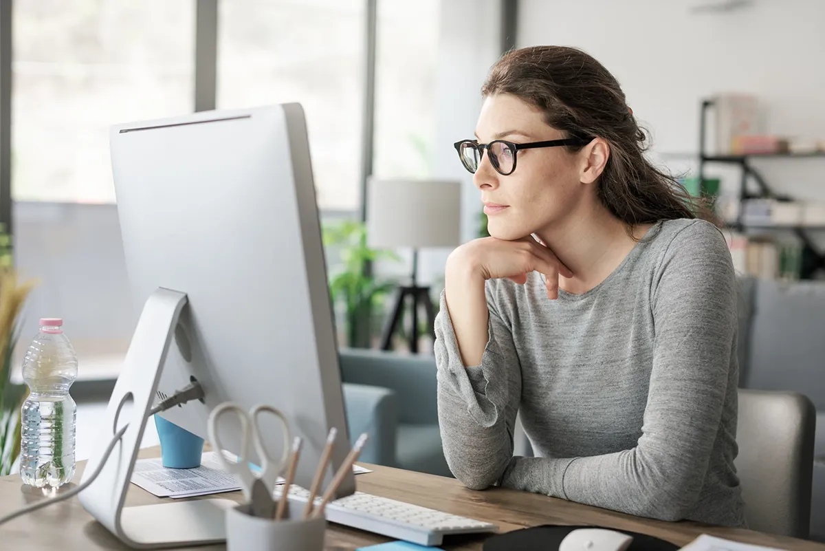 A woman wearing glasses leans forward to look at her computer.